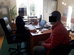 Na zdjęciu widoczny jest policjant nieumundurowany siedzący przy biurku, Obok siedzi na krześle zatrzymany mężczyzna ubrany w kurtkę koloru czerwonego.