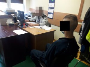 Na zdjęciu widoczny nieumundurowany policjant siedzący przy biurku w pomieszczeniu służbowym. Policjant przesłuchuje zatrzymanego mężczyznę, który siedzi obok na krześle i ma założone kajdanki.