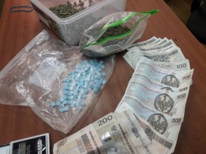 Na zdjęciu leżą na biurku w pomieszczeniu służbowym zabezpieczone przez policjantów narkotyki w postaci niebieskich tabletek i marihuana w kolorze zielonym, waga elektroniczna i banknoty.