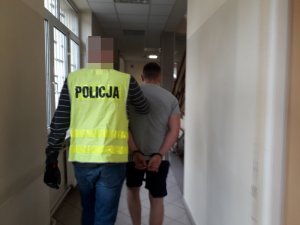 Na zdjęciu zatrzymany mężczyzna w kajdankach. Jest prowadzony korytarzem w jednostce Policji przez nieumundurowanego policjantka, który ma żółtą kamizelkę z napisem Policja.