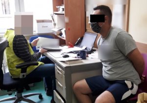Na zdjęciu siedzi na krześle w pomieszczeniu służbowym zatrzymany mężczyzna, ubrany w krótkie niebieskie spodenki i koszulkę koloru szarego. Obok przy biurku siedzi nieumundurowany policjant w  żółtej kamizelce z napisem Policja.