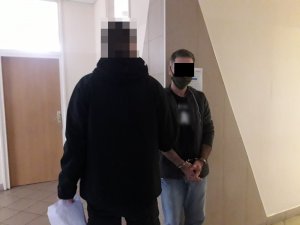 Na zdjęciu widoczny zatrzymany mężczyzna, który ma założone na ręce kajdanki. Z rękę trzyma go nieumundurowany policjant.