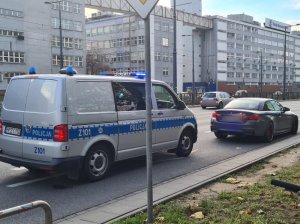 Na zdjęciu widoczne poszukiwane auto marki BMW i policyjny oznakowany radiowóz. Auta stoją na wzdłuż ulicy.