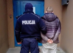 Na zdjęciu widoczny umundurowany policjant, który trzyma z rękę zatrzymanego. Obaj stoją przed drzwiami budynku komisariatu.