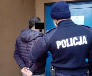 Na zdjęciu widoczny umundurowany policjant, który trzyma z rękę zatrzymanego. Obaj stoją przed drzwiami budynku komisariatu.