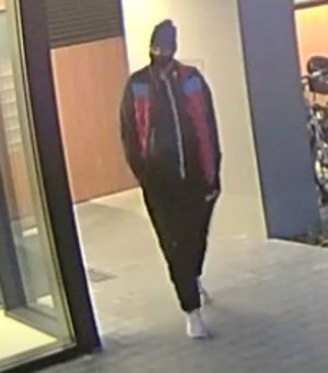 Na zdjęciu widoczny poszukiwany mężczyzna. Jest on ubrany w kurtkę koloru czarno-czerwonego, ma czapkę na głowie oraz spodnie dresowe koloru czarnego. Buty białe sportowe.