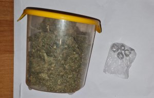 Na zdjęciu widoczny plastikowy pojemnik z zawartością suszu roślinnego koloru zielonego. W torebce foliowej widać zawiniątka aluminiowe z zawartością amfetaminy.