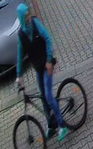 Naz zdjęciu widoczny mężczyzna trzymający rower, ubrany w kurtkę koloru czarno-niebieskiego, spodnie jeansowe. Na głowie ma założony kaptur koloru niebieskiego.