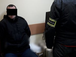 Na zdjęciu widoczny zatrzymany mężczyzna siedzący na krześle w pokoju służbowym. Obok stoi nieumundurowany policjant.