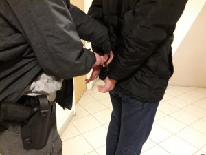 Na zdjęciu widoczny zatrzymany mężczyzna, który ma nakładane na ręce kajdanki przez nieumundurowanego policjanta.