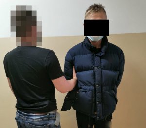 Na zdjęciu widoczny nieumundurowany policjant, który za rękę trzyma zatrzymanego mężczyznę ubranego w kurtkę koloru granatowego i spodnie sportowe koloru czarnego.