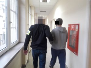 Na zdjęciu widoczny nieumundurowany policjant, który prowadzi po korytarzu służbowym zatrzymanego mężczyznę.