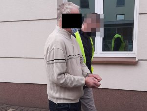 Na zdjęciu widoczny nieumundurowany policjant w żółtej kamizelce, który prowadzi zatrzymanego mężczyznę.