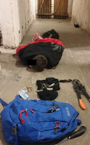 Na zdjęciu widoczny zatrzymany mężczyzna, który posiada założone kajdanki na ręce, leży na podłodze w piwnicy. Obok leżą narzędzia jak obcęgi, noże i plecak koloru niebieskiego.
