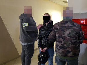 Na zdjęciu widoczni nieumundurowani policjanci, którzy trzymają za rękę zatrzymana kobietę.