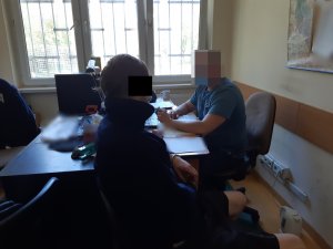 Na zdjęciu widoczny zatrzymany mężczyzna, który siedzi na krześle, obok przy biurku siedzi nieumundurowany policjant.