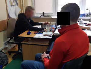 Na zdjęciu widoczny nieumundurowany policjant, który siedzi przy biurku. Obok siedzi zatrzymany mężczyzna.