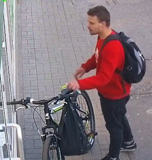 Na zdjęciu widoczny mężczyzna ubrany w bluzę sportową koloru czerwonego, spodnie ciemne. Na plecach ma założony plecak, trzyma rower.