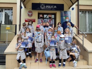 Na zdjęciu widoczne zuchy i umundurowany policjant przed wejściem do budynku komendy.