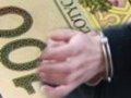 Na zdjęciu widoczny banknot oraz dłonie zatrzymanego w kajdankach.