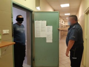 Na zdjęciu widoczny umundurowany policjant, w drzwiach pomieszczenia dla osób zatrzymanych stoi zatrzymany mężczyzna.