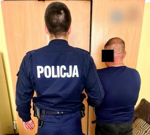 Na zdjęciu widoczny umundurowany policjant, przy nim stoi zatrzymany mężczyzna.