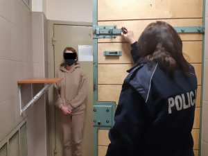 Na zdjęciu widoczna umundurowana policjantka, który stoi przy zatrzymanej kobiecie w pomieszczeniu dla osób zatrzymanych.