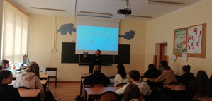 Na zdjęciu widoczny umundurowany policjant oraz uczniowie liceum, którzy siedzą w ławkach szkolnych.