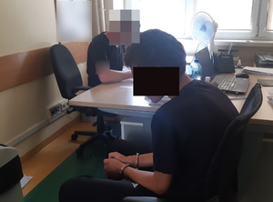 Policjant siedzi przy biurku, zatrzymany siedzi na krześle obok.