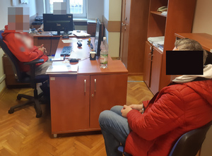 Zatrzymany siedzi na krześle, obok przy biurku siedzi policjant.